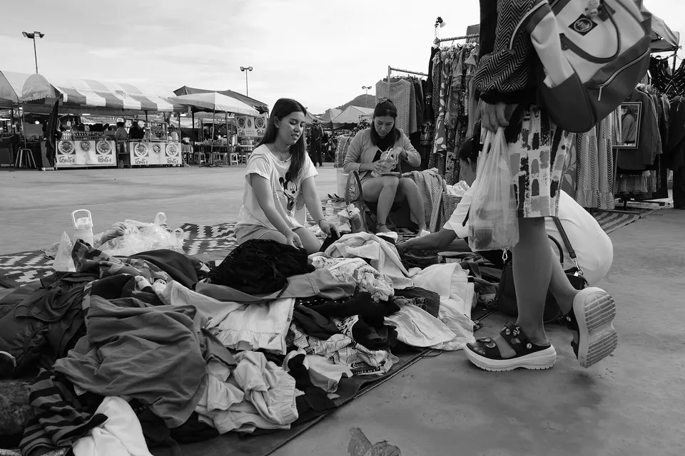 Shoppers sifting through a large pile of secondhand clothes on the ground at an open-air market, with stalls and umbrellas behind